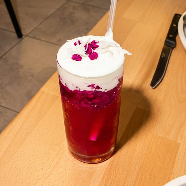 Vibrant pink drink with white foam, coconut flakes, and dried flower petals on a wooden table.
