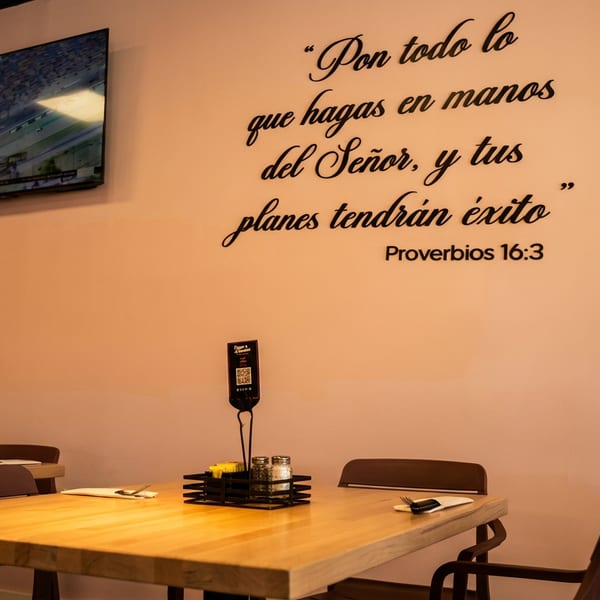 Restaurant dining area with light wood tables, brown chairs, and a Spanish scripture quote on the wall.
